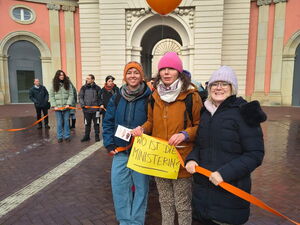 Frauen halten ein orangenes Band. 3 Frauen im Vordergrund mit orangenem Luftballon und einem Schild in den Händen. Text: Wo ist die Ministerin?