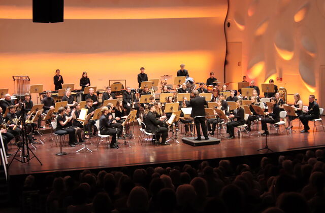 Gesamte Bühne, gesamtes Orchester. Dirigent in der Mitte mit Blick zum Orchester. Viele Frauen und Männer mit Blasinstrumenten, Streichinstrumenten und ein Schlagzeug. Die Bühne ist sanft beleuchtet.