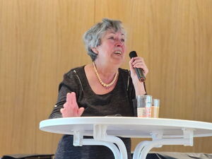 Frau mit Mikrofon steht an einem runden weißen Stehtisch mit Getränken und spricht.