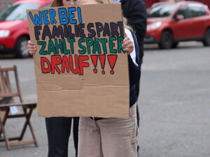 Eine Person hält eine beschriebene Pappe hoch. Auf dem Schild steht: "Wer bei Familien spart, zahlt später drauf".