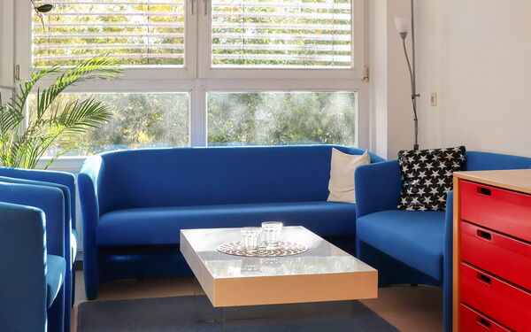 Sprechzimmer. 2 blaue Sessel, 2 Blaue Sofas und ein rechteckiger Tisch in der Mitte.