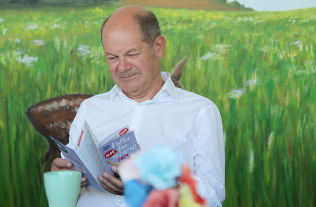 Mann sitzt vor einem Wandbild, dass eine weite Wiese mit Blumen zeigt. Mann guckt sich den Flyer von Kultur für Jeden an. Der Mann ist älter, hat eine Halbglatze, Haare an den Seiten sind kurz und grau. Er trägt ein weißes Hemd vor ihm eine Tasse.