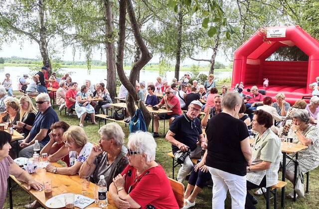 Viele Menschen an Biertischgarnituren. Essen und Getränke auf den Tischen. Im Hintergrund eine rote Hüpfburg.