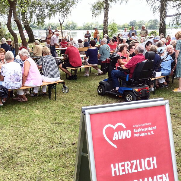Viele Menschen an Biertischgarnituren. Essen und Getränke auf den Tischen. Im Hintergrund ein Gewässer.