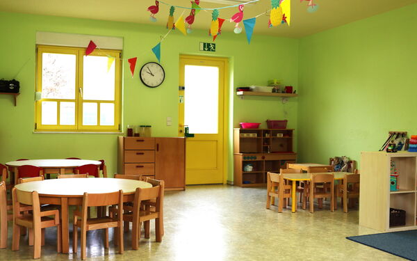 Spielraum. Zwei runde kleine Tische mit kleinen Stühlen, ein rechteckiger kleiner Tisch mit kleinen Stühlen, Regale, ein Schrank. An der Decke hängen Wimpel und eine Girlande mit Ananas und Flamingos.