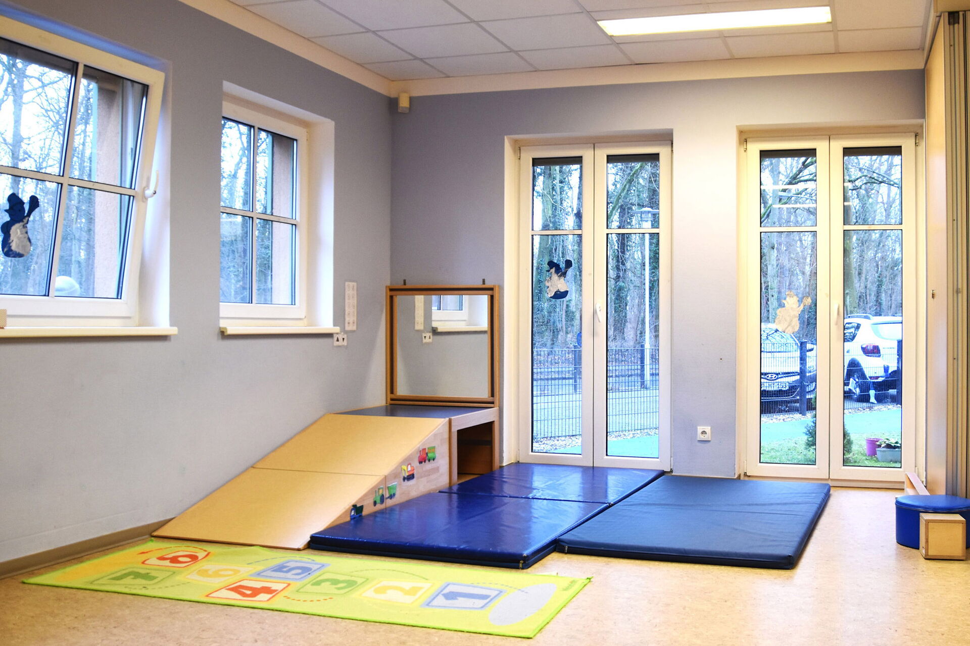 Sportraum, große blaue Matten auf dem Boden, links eine Schräge die zu einem klein Podest mit Wandspiegel in der Ecke führt.