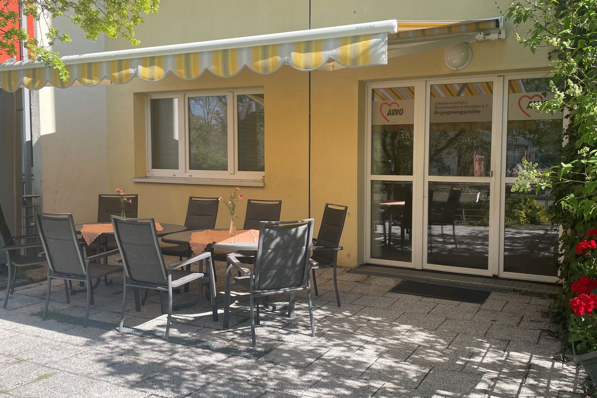 Terrasse mit Stühlen und Tischen. Eine Markise spendet Schatten, die Blumenkästen sind mit Geranien bepflanzt.
