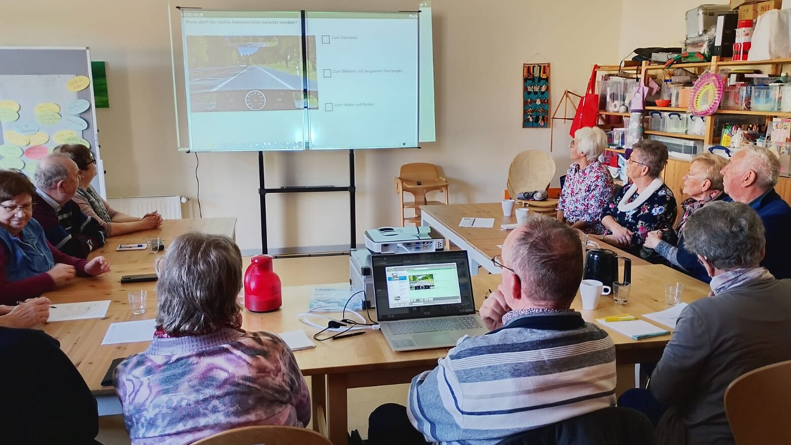 11 Senior*innen sitzen im Raum "Familiencafé" zusammen am Tisch. An der Wand hängt eine Tafel, auf der ein Bild aus einer Fahrschulprüfung zu shene ist.