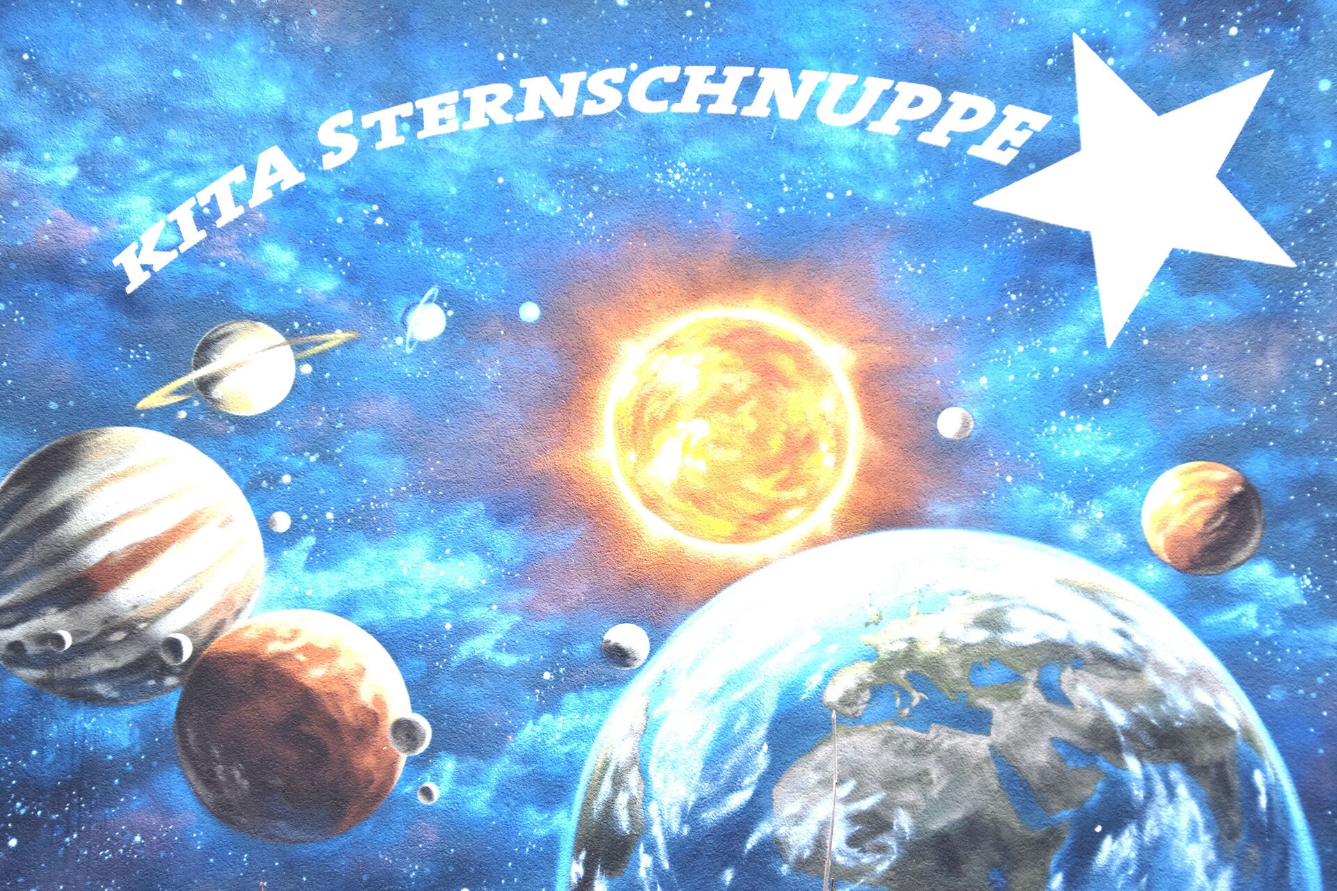eine Darstellung unseres Sonnensystems mit Sonne, Erde und mehreren Planeten. Im Hintergrund ist ein Sternenhimmel zu sehen. Oben im Bild steht in großen weißen Buchstaben "Kita Sternschnuppe". Es wirkt wie ein Wandbild