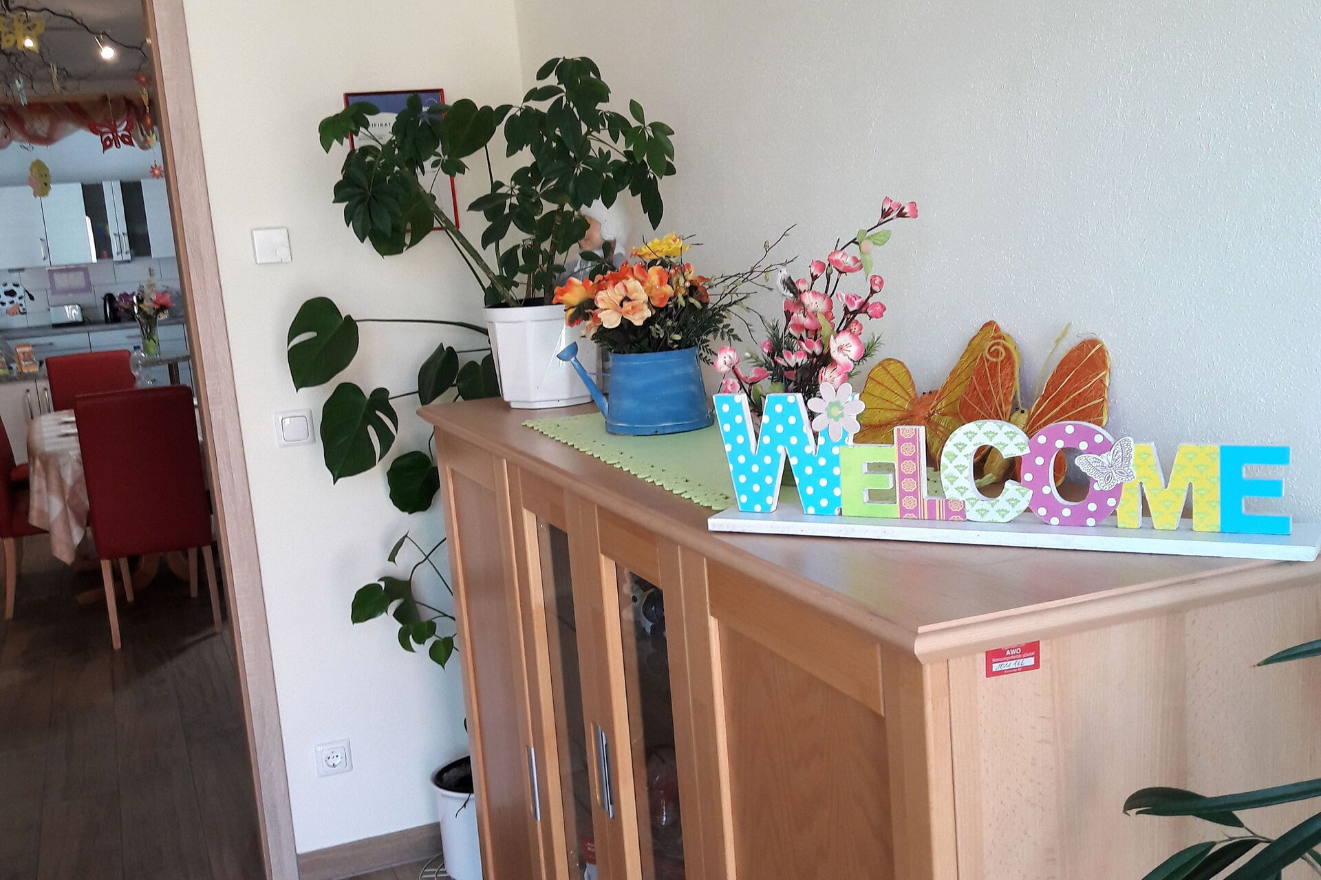 Flur. Eine Kommode an der Wand rechts. Darauf Blumen und ein buntes Schild: WELCOME.
