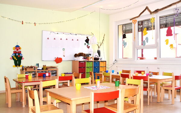 helleR, freundlicher Kindergartenraum. Im Vordergrund stehen mehrere kleine Tische mit bunten Kinderstühlen. An der Wand befindet sich eine große weiße Tafel, und ein farbenfroher Clown ist aufgehängt