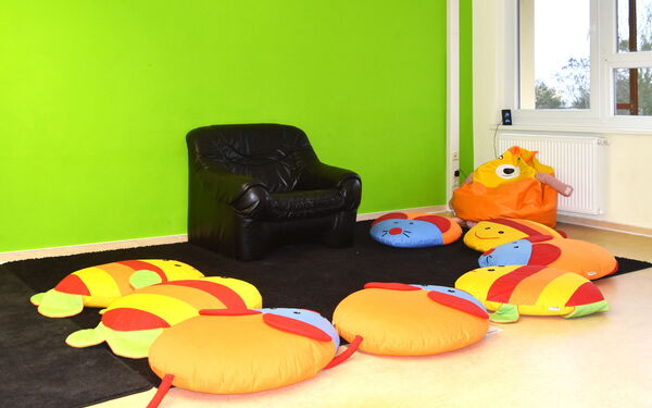 Zimmer mit einer grünen Wand. An der Wand steht ein schwarzer Sessel. Um den Sessel liegenn 8 bunte Sitzkissen, die aussehen wie Mäuse und Fische.