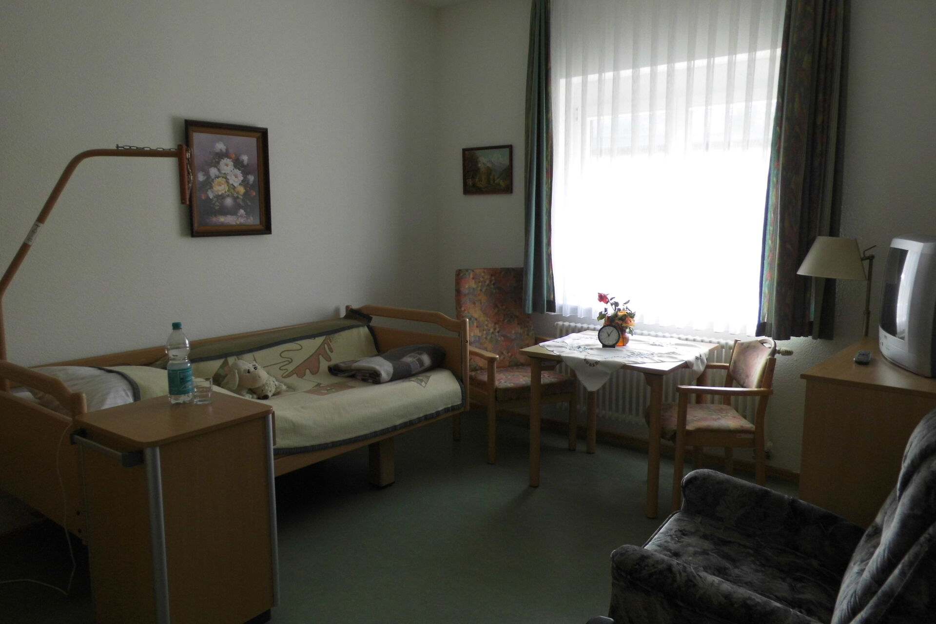 Bewohnerzommer, gemütlich eingerichtet mit Bett, Sessel, Tisch und 2 Stühlen am Fenster.