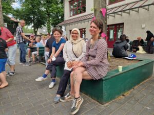 Draußen, drei Frauen sitzen unter einem Baum, und lächeln in die Kamera. Drumherum ist frohes treiben. Kinder sitzen auf Bänken.