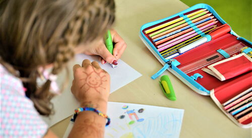 Mädchen sitzt am Tisch und schreibt mit der linken Hand. Auf dem Tisch liegen eine Federmappe zum aufklappen mit Buntstiften, Linial und Anspitzer. Und ein gemaltes Bild liegt unter dem rechten Arm des Mädchens.