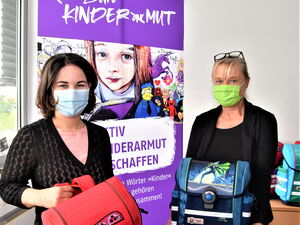 Im Büro Kinderarmut: Zwei Frauen mit Mund-Nasen-Schutz Masken. Jede hat einen Schulranzen in den Händen. Baerbock hat einen roten Ranzen, Schweers einen blauen Ranzen.