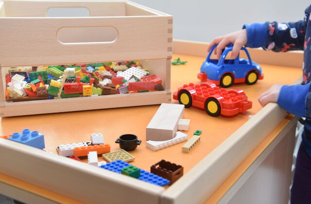 Spieletisch mit Lego und keinem blauen Auto. Eine Kinderhand umfasst die Kante des Tisches mit der anderen Hand schiebt es das Auto.