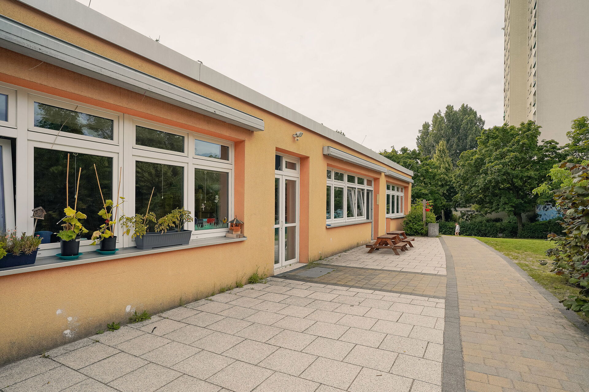 Flaches langes Gebäude mit orangener Fassade. Auf dem Fensterbrett stehen Pflanzen.