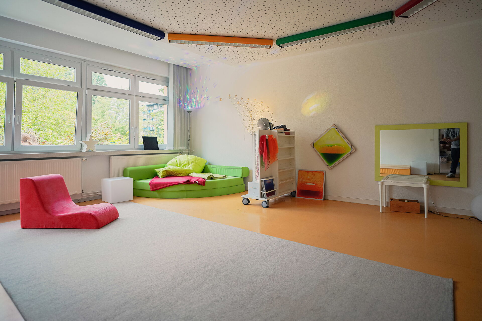 Großer heller Raum, große Fenster lassen Licht hinein. Eine grüne Sitzecke, kleines Ragal mit Rollen als Raumtrenner und ein Spiegel mit grünem Rahmen. Ein hellgrauer Teppich liegt mittig im Raum.