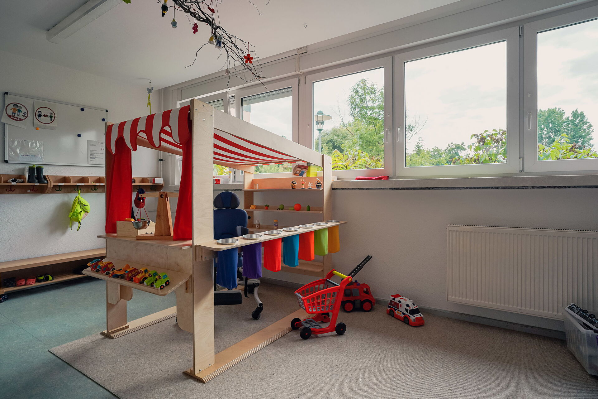 Helles Spielzimmer, mit Kinder Spielzeug, Krankenwagen, Feuerwehr und ein Einkaufsladen für Kinder aus Holz mit rot-weiß gestreifter Markise.