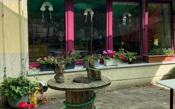 Gebäude mit vielen Fenstern, auf der Fensterbank sind bepflanzte Blumenkästen. Davor steht ein runder Holztisch. An drei der Fenster sind Regenbogenbilder.