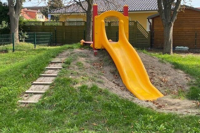 Eine gelbe Rutsche auf eine kleinen Hügel. Wiese, drei Bäume.