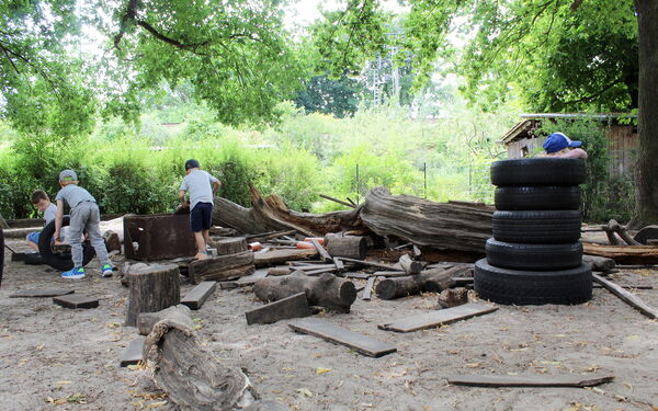 Holz-Baustelle im Garten. Reifen sind aufgestapelt und Holzstücke, Holzstämme liegen rum. 4 Kinder spielen dort.