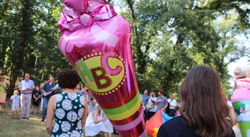 Großer Luftballon in Form einer Zuckertüte pink mit A-B-C Bild. Einige Menschen draußen auf einer Wiese mit Bäumen.