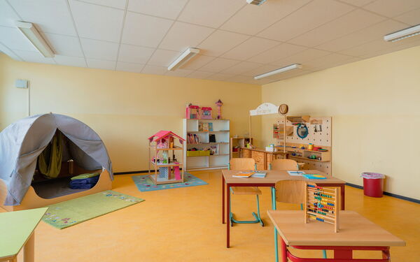 Spielzimmer, auf einem kleinen Tisch steht ein Rechenschieber. Ein Puppenhaus steht auf einem Kinderteppich, ein Regal an der Wand mit Spielzeug: