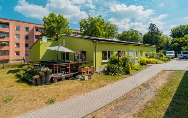 Grüner Flachbau mit einer Terrasse mit Sonnenschirm und Bepflanzung.