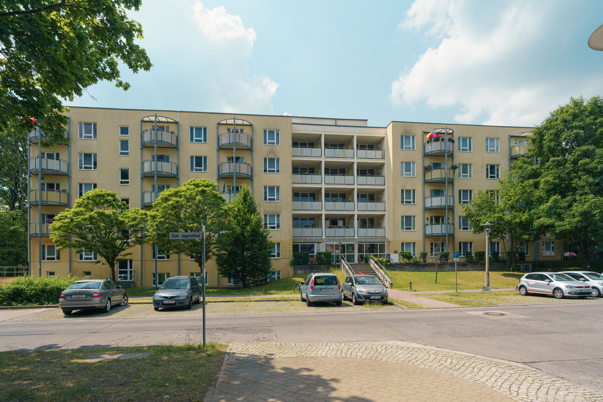 Eine Straße, wenige parkender Autos. Dahinter und großes Wohnhaus mit gelber Fassade und Balkonen.