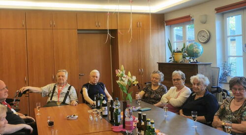 Alte Frauen und Männer an einem großen Tisch. Auf dem Tisch stehen Getränke und Gläser.