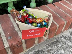 Draußen an einem Gartenzaun. Osternest mit Schokoladeneiern und anderen Süßigkeiten. Daran in ein AWO Schildchen gebunden. Text: Bunte Grüße