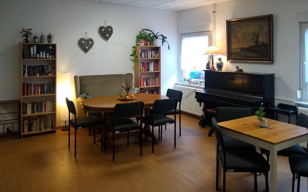 Dunkler Raum, mit Tischen, Stühlen und Sofa. 2 Bücherregale stehen an der Wand.