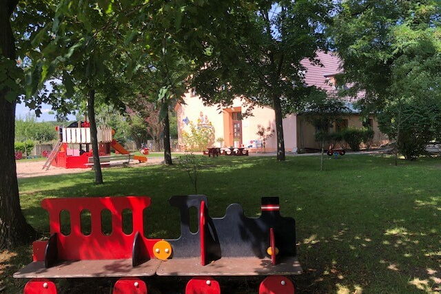 Eine Sitzbank für Kinder rot-schwarz. Sieht aus wie eine Eisennbahn. Die Lock ist schwarz. In Hintergrund Spielplatz, Sandkasten und ein Haus.