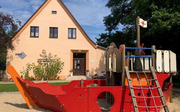 Spielplatz, Klettergerüst als Schiff mit Fahne und Steuerrad und Bullauge. Dahinter ein Haus mit Spitzdach. Das Kita-Gebäude.