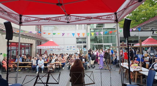 Viele Menschen auf einem Innenhof. Es gibt Biertischgarnituren, AWO Pavillons. Eine Bühne mit Lautsprechern und Notenständern. Die Menschen sehen zur Bühne.