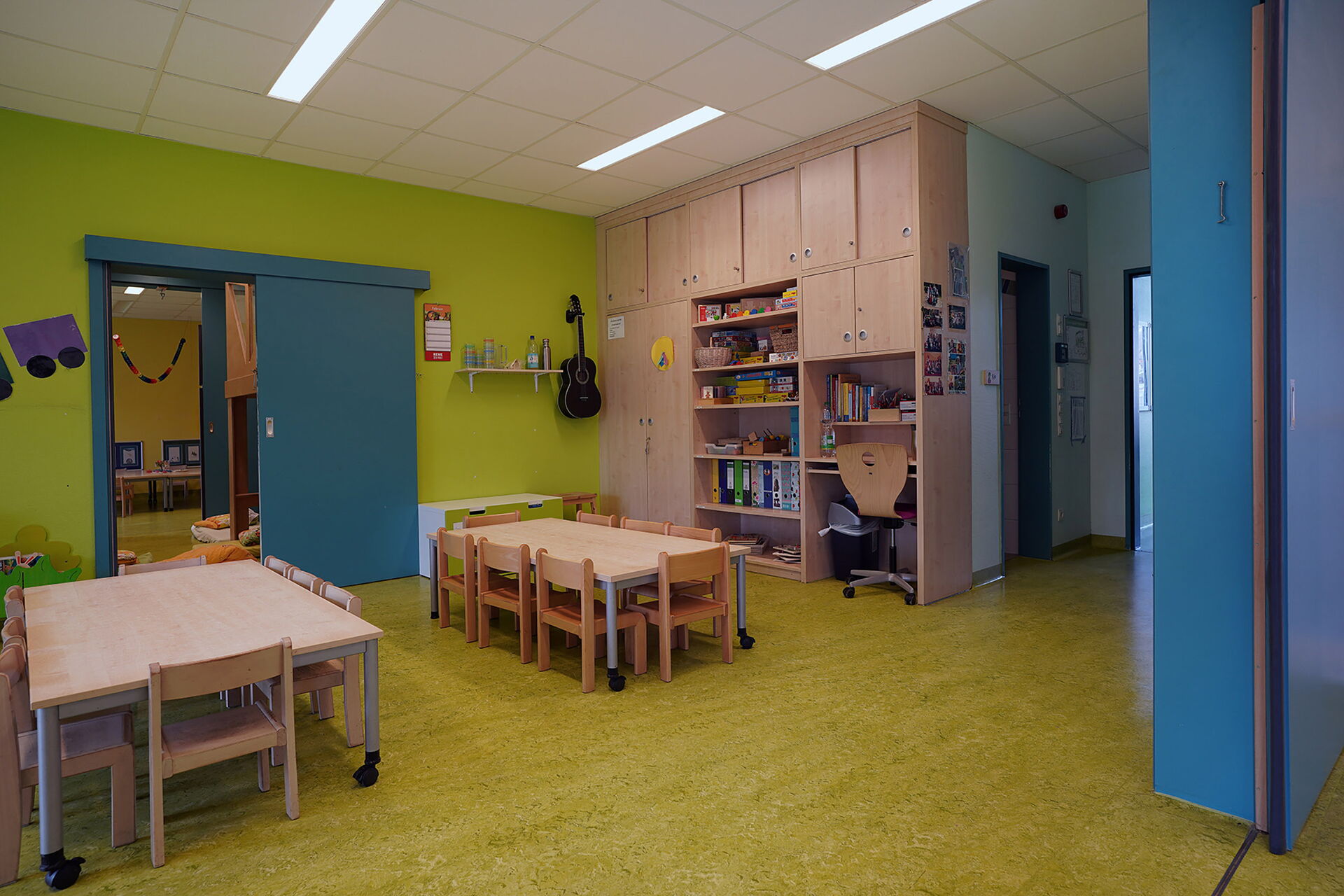 Ein Kindergartenraum mit Tischen und Stühlen, einer Wand und Fußboden in grüner Farbe. Regale und Schränke.