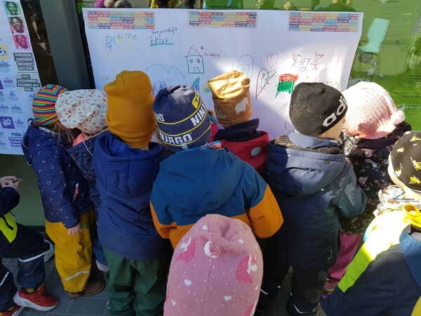Kleine Kinder draußen mit Jacken und Mützen, Sie stehen alle mit Rücken zur Kamera vor einem Plakat mit Kinder-Zeichnungen.