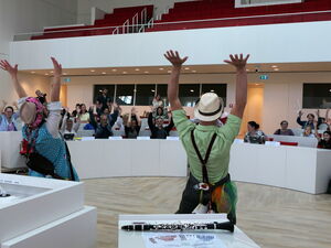 Klinikclowns sorgen für gute Laune im weißen Plenarsaal. 2 Clowns strecken die Arme nach Oben, Kinder und Erwachsene auf den Sitzplätzen machen mit.