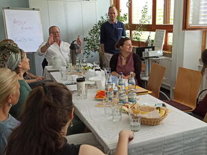 Heller Raum, Menschen sitzen an einem Tisch mit Wasserflaschen und Gläsern. Bei einem Mann an der Stirnseite steht ein Flipchart beschriftet mit: Herzlich willkommen.