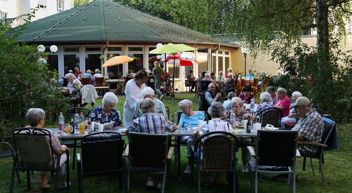 Draußen, Tische sind aneinander gereiht. Ältere Menschen sitzen essen und trinken. Ein großer Baum spendet Schatten, sonst sind kleine farbige Sonnenschirme aufgestellt.