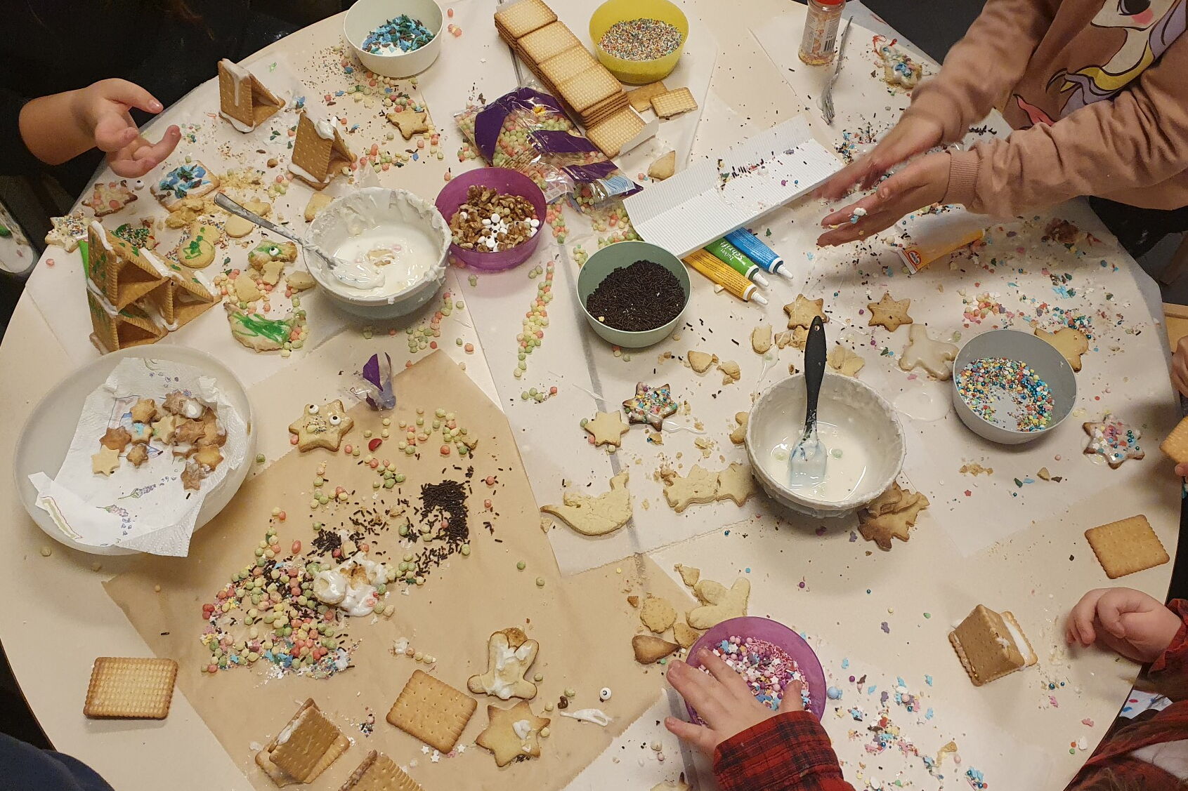 Weihnachtsplätzchen dekorieren. Runder Tisch von oben. Plätzchen, Glasuren, bunte Streusel. Kinderhände die mit dekorieren beschäftigt sind.