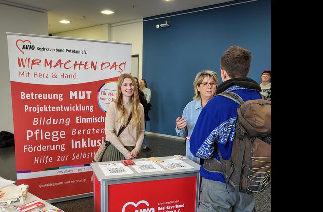 Ein AWO Stand auf einer Messe Mit Info- Material. Eine junge Frau steht hinter dem Stand und lächelt in die Kamera. Eine weitere Frau ist im Gespräch mit einem Mann.
