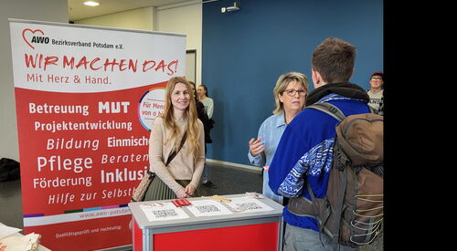 Ein AWO Stand auf einer Messe Mit Info- Material. Eine junge Frau steht hinter dem Stand und lächelt in die Kamera. Eine weitere Frau ist im Gespräch mit einem Mann.