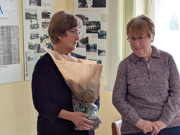 2 ältere Frauen. Die links hält ein Glas mit einem Blumenstrauß der in Papier gewickelt ist. Die andere Frau lächelt.