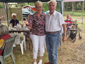 Eine Frau und ein älterer Mann mit weißen Haaren und weißem kurzärmligen Hemd stehen nebeneinander und lächeln in die Kamera. Dahinter Menschen an Kaffeetischen.