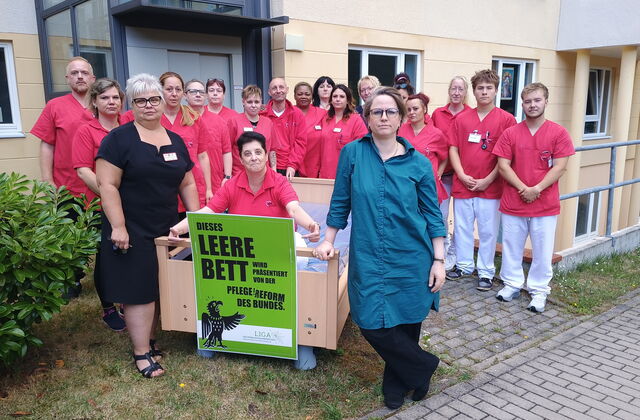Viele Pflegende in Arbeitskleidung vor einer Einrichtung. Ein Pflegebett steht vor ihnen, darin sitzt eine Pflegerin und zeigt auf ein angebrachtes grünes Schild.
