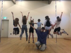 Raum / Tanzstudio. 6 Mädchen von hinten im Sprung fotografiert. Ein Junge kniet auf dem Boden und breitet die Hände nach oben aus.
