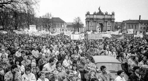 Schwarz-Weiß Foto: Vor dem Brandenburger Tor in Potsdam stehen dicht gedrängt ganz viele Menschen. Einige von ihnen halten Plakate hoch, auf denen sie Freiheit oder das Absetzen der Regierung fordern.